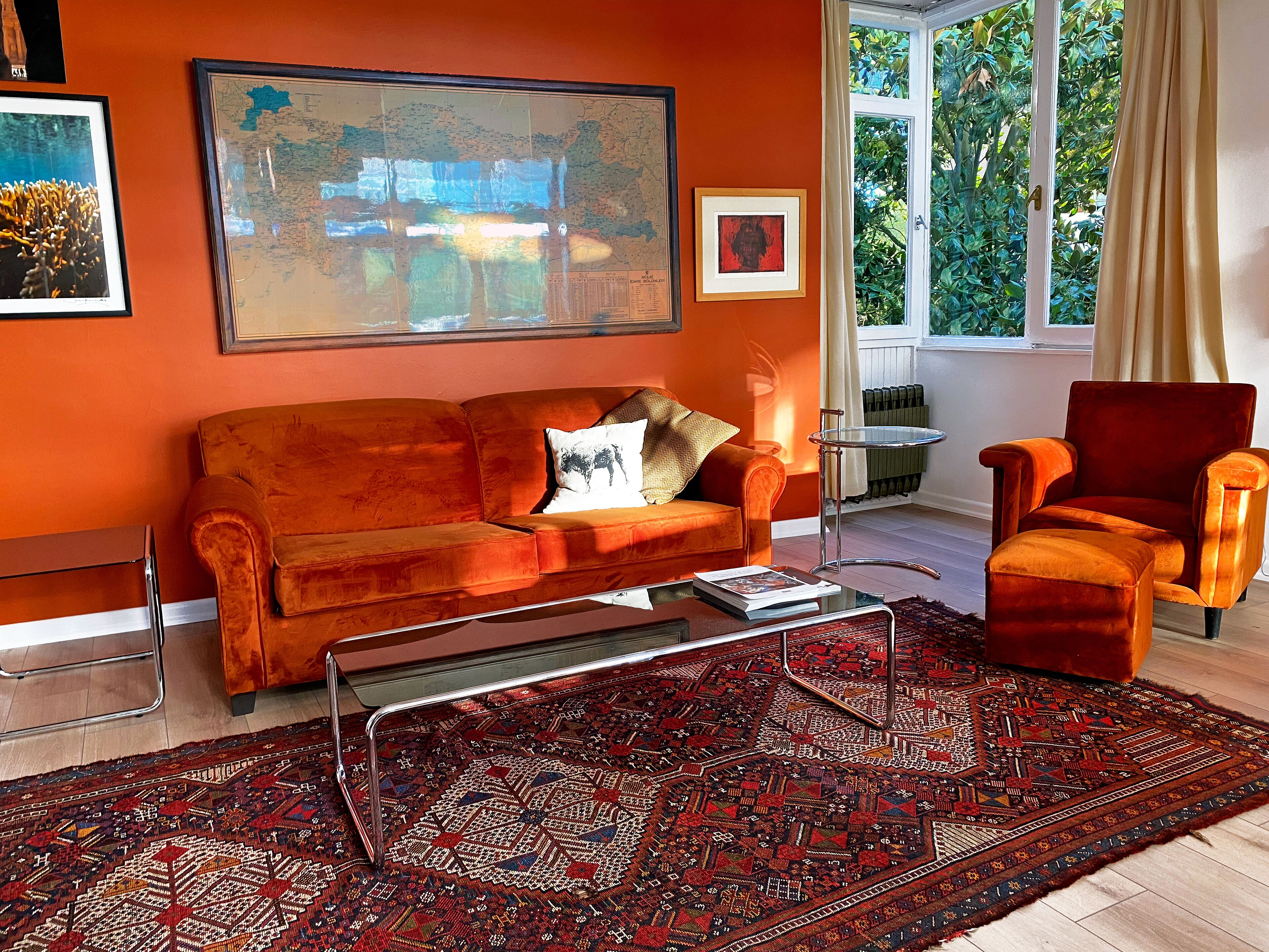 Una habitación con un sofá naranja, un sillón rojo, una mesa de centro de vidrio y una alfombra estampada.