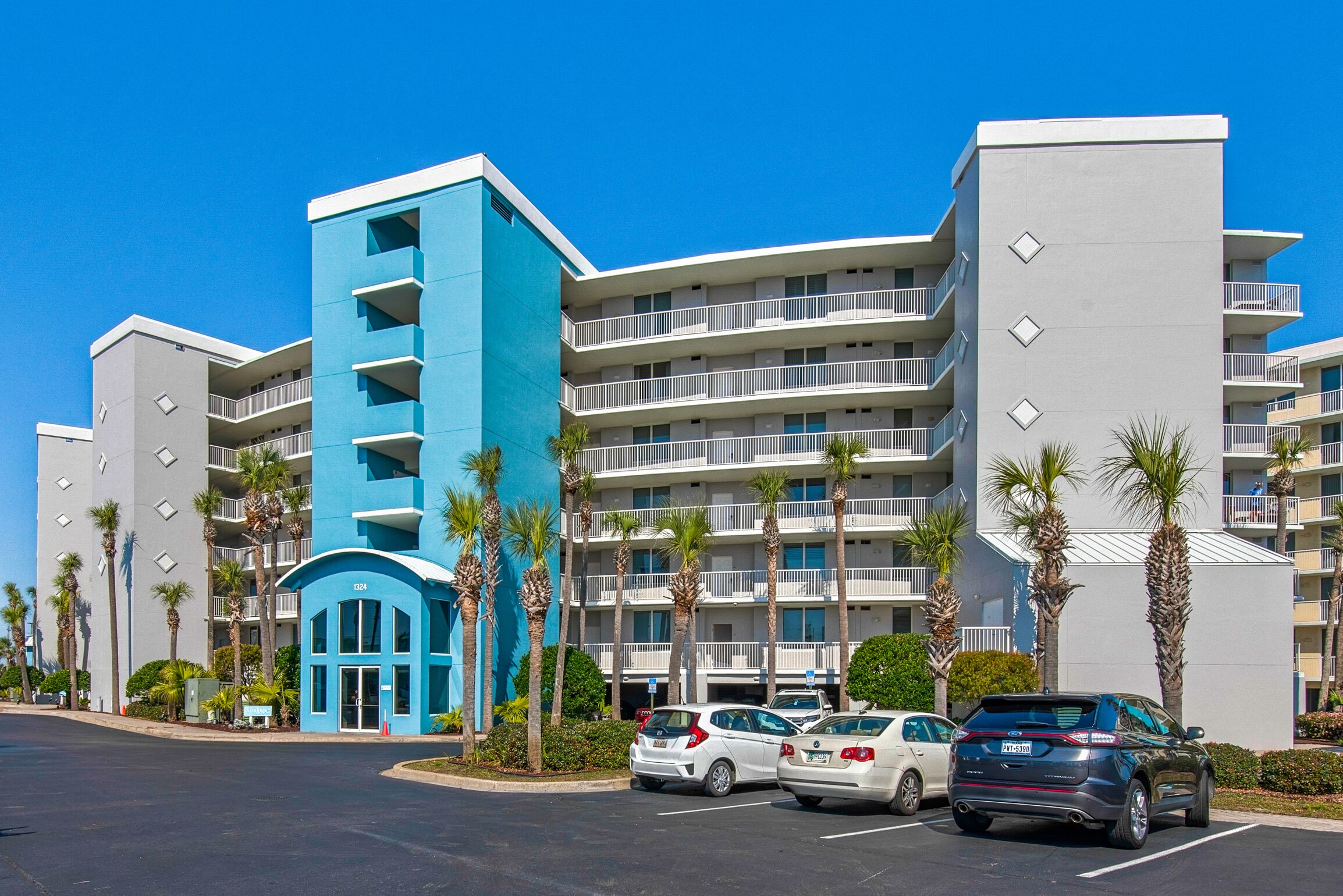 Un hôtel à plusieurs étages, avec une façade bleue et blanche, un parking et des palmiers.