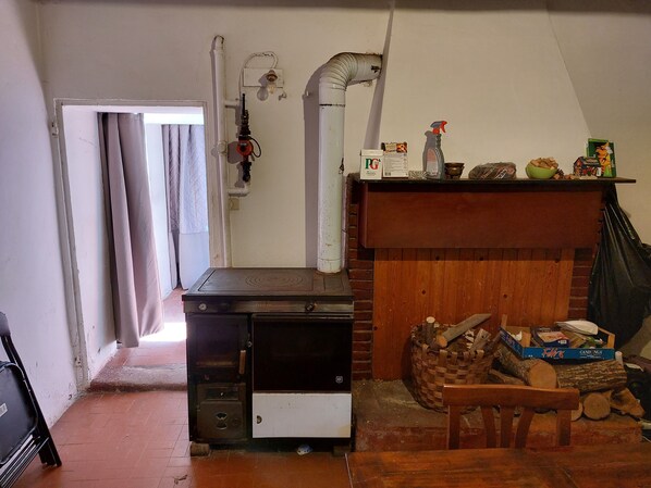 A kitchen with a stove, a sink, and a wooden shelf above the stove. There is a basket of firewood and a chair in the corner. - Remarkable 3-bed Cottage in Tortola (Predappio)