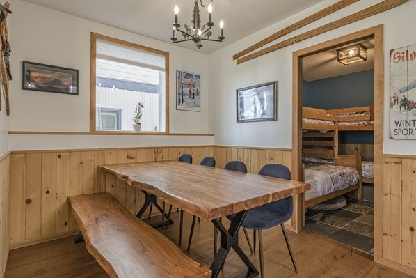 Una mesa de comedor de madera con bancos, una araña, cuadros enmarcados y una zona con literas.