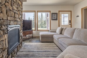 Una sala de estar con chimenea de piedra, televisor, sofá seccional color beige y ventanas amplias con vista a árboles.