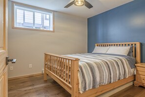Un dormitorio con una cama de madera, una mesita de noche, un ventilador de techo, una ventana y una pared azul.