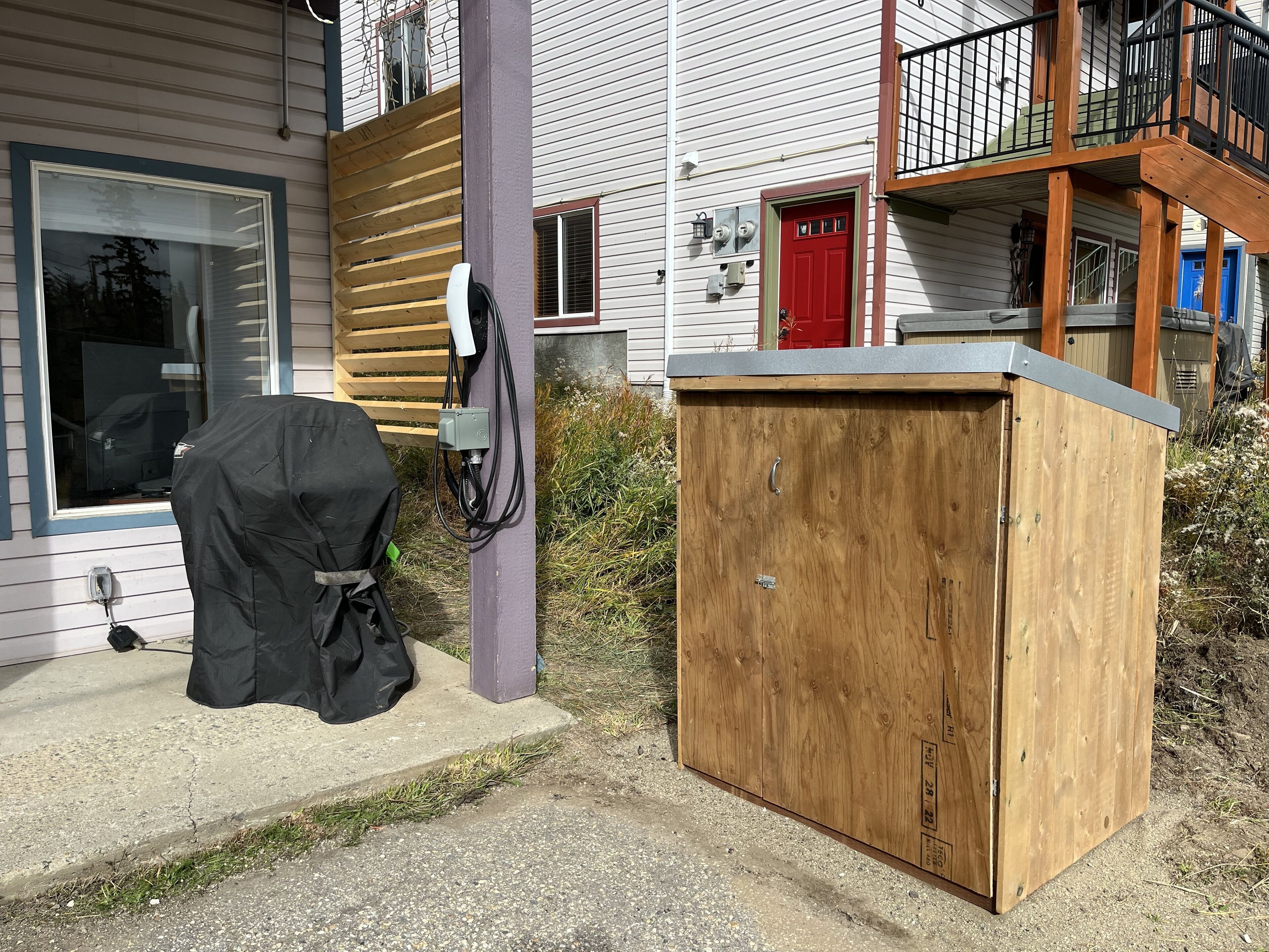 Un petit abri en bois, servant d’espace de rangement, se trouve à côté d’une maison avec une porte rouge et un balcon.