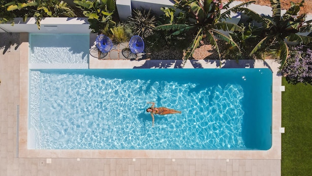 Vista aérea de una piscina rectangular con agua azul cristalina, rodeada por un solárium con baldosas y abundante vegetación.