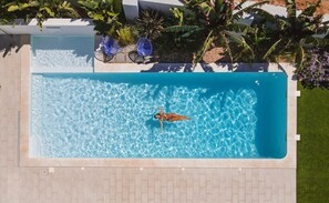 Vue aérienne d’une piscine rectangulaire à l’eau d’un bleu limpide, entourée d’une terrasse carrelée et d’une végétation luxuriante.