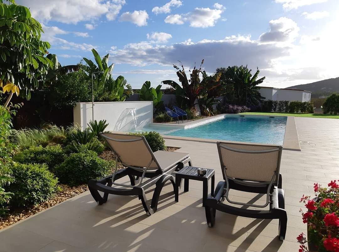 Un espace piscine agrémenté de chaises longues et d’une petite table, entouré d’une végétation luxuriante et sous un ciel dégagé.