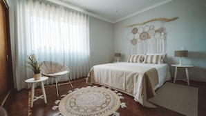 Un dormitorio con una cama, dos mesitas de noche, una silla, un adorno en la pared y un amplio ventanal con cortinas translúcidas.