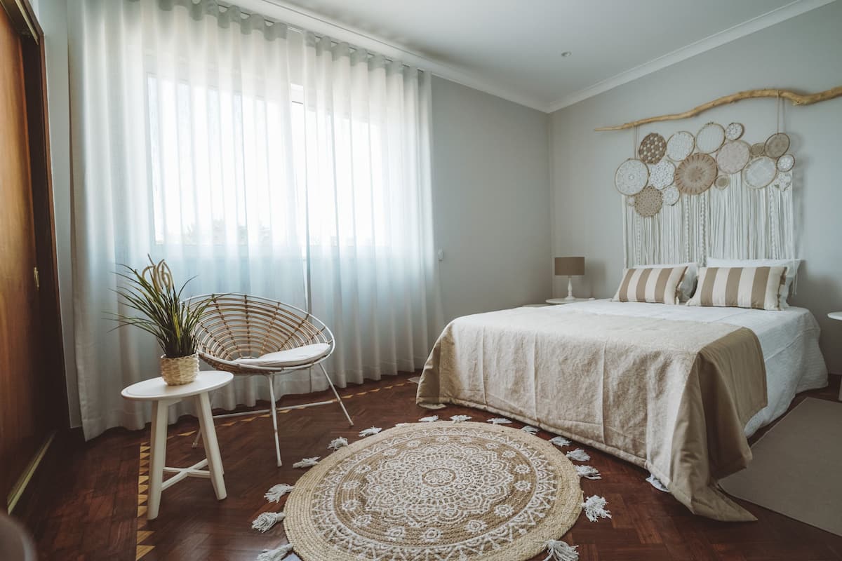 Ein Schlafzimmer mit einem Bett, zwei Nachttischen, einem Sessel, einem Wandteppich und einem großen Fenster mit durchsichtigen Vorhängen.