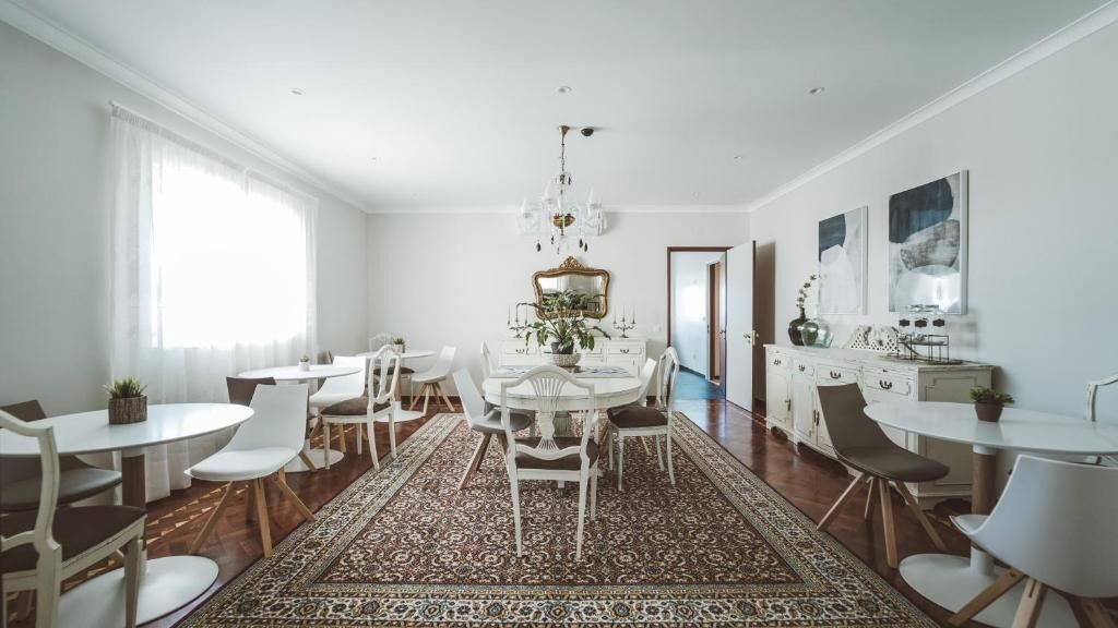 Uma sala de jantar espaçosa com uma grande mesa redonda cercada por cadeiras brancas, um tapete estampado e um candelabro.