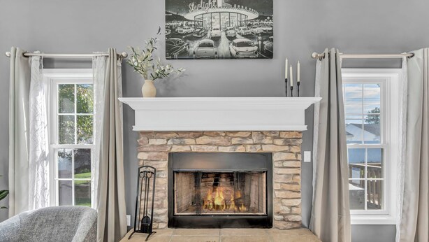 Une cheminée en pierre avec un manteau de couleur blanche, encadrée de deux fenêtres ornées de rideaux, un mur gris et un tableau encadré au-dessus.