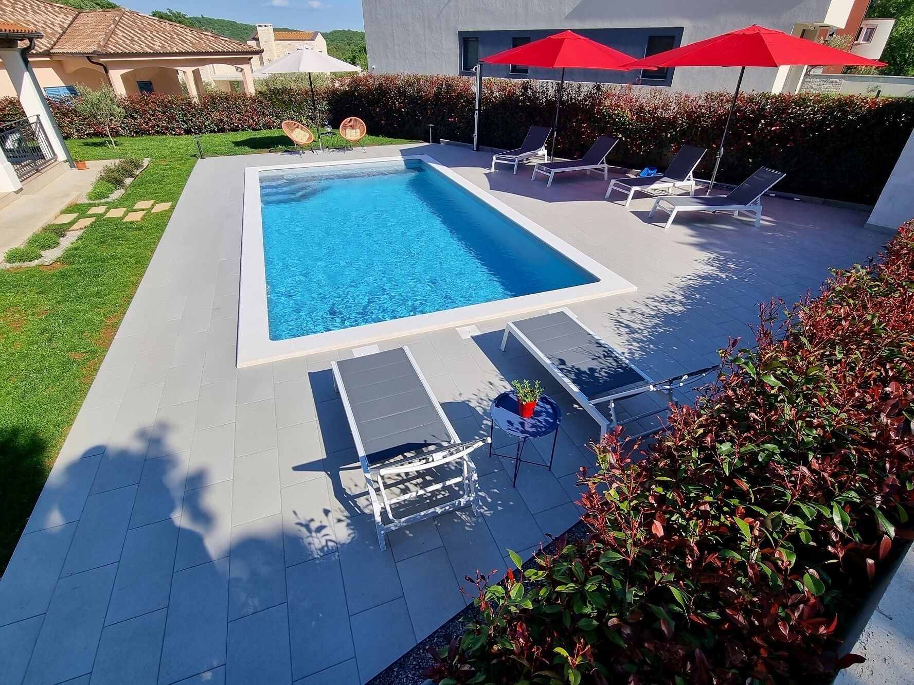 Una piscina rectangular con sillas de descanso y sombrillas, rodeada de césped y setos cuidados.