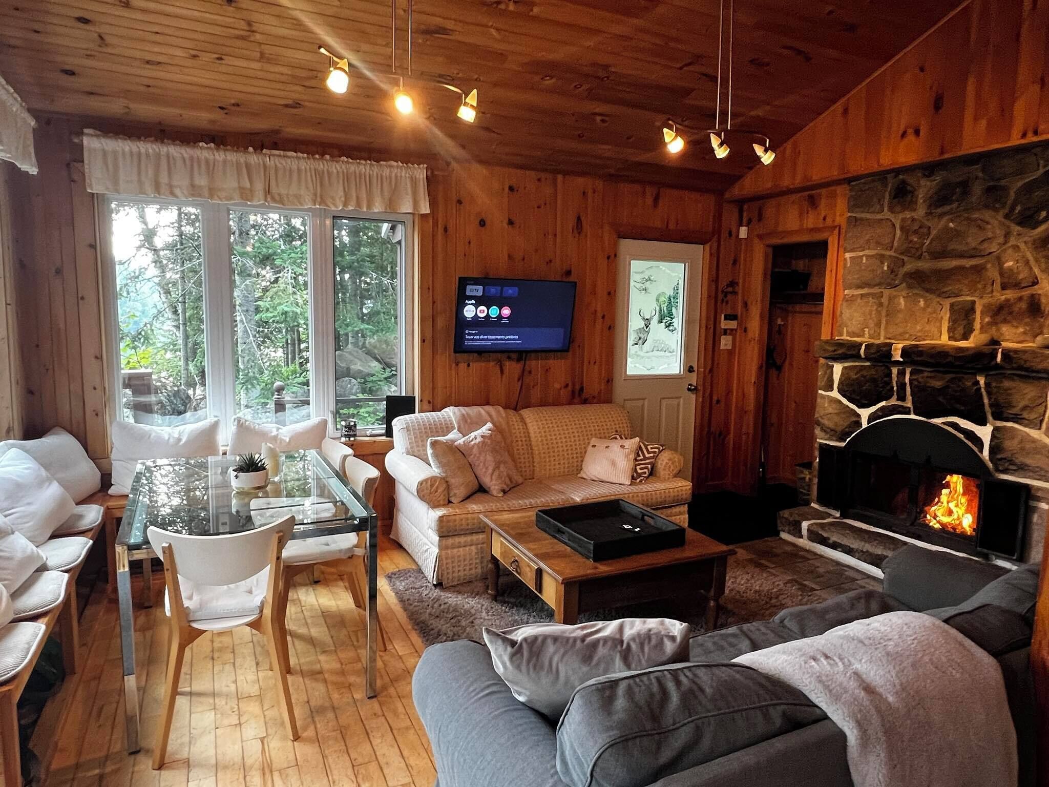 L’intérieur chaleureux d’un chalet avec une cheminée en pierre, des murs en bois, un téléviseur et un coin repas comprenant une table et des chaises en verre.
