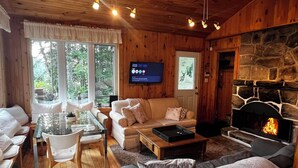 L’intérieur chaleureux d’un chalet avec une cheminée en pierre, des murs en bois, un téléviseur et un coin repas comprenant une table et des chaises en verre.