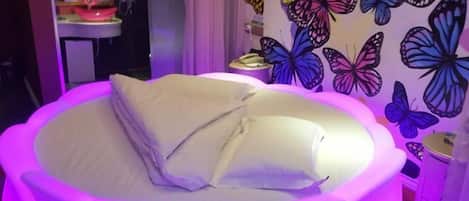Una cama con colchón y almohadas blancos, rodeada por un cabecero rosa iluminado.