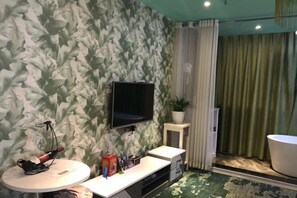 A room with a patterned wallpaper, a mounted TV, a small table with a plant, and a bathroom with a green curtain. - Yimengshu Hotel (Lichuan)