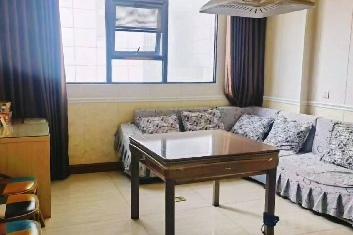 Una sala de estar con sofá, una mesa de centro de vidrio, una mesita auxiliar de madera y una ventana con cortinas.