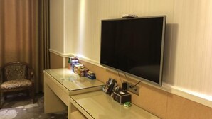 A hotel room with a flat-screen TV mounted on the wall, a glass desk with various items, a patterned chair, and a curtain. - Yimengshu Hotel (Lichuan)