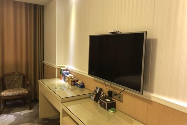 A hotel room with a flat-screen TV mounted on the wall, a glass desk with various items, a patterned chair, and a curtain. - Yimengshu Hotel (Lichuan)