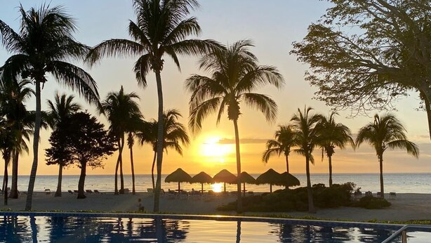 Una piscina amb vistes a la posta de sol, palmeres i paraigües de platja.