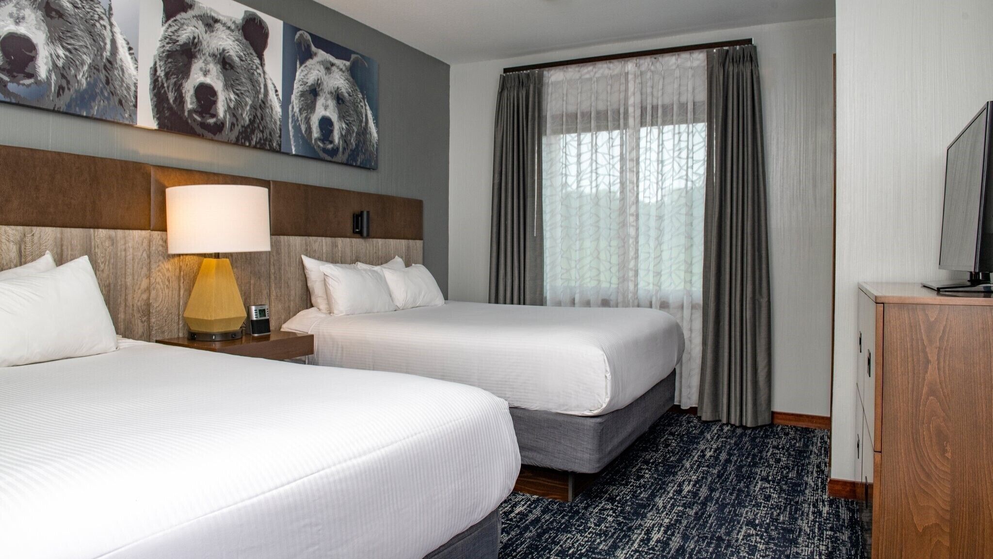 A hotel room with two beds, a lamp, a TV, and a large window with curtains.