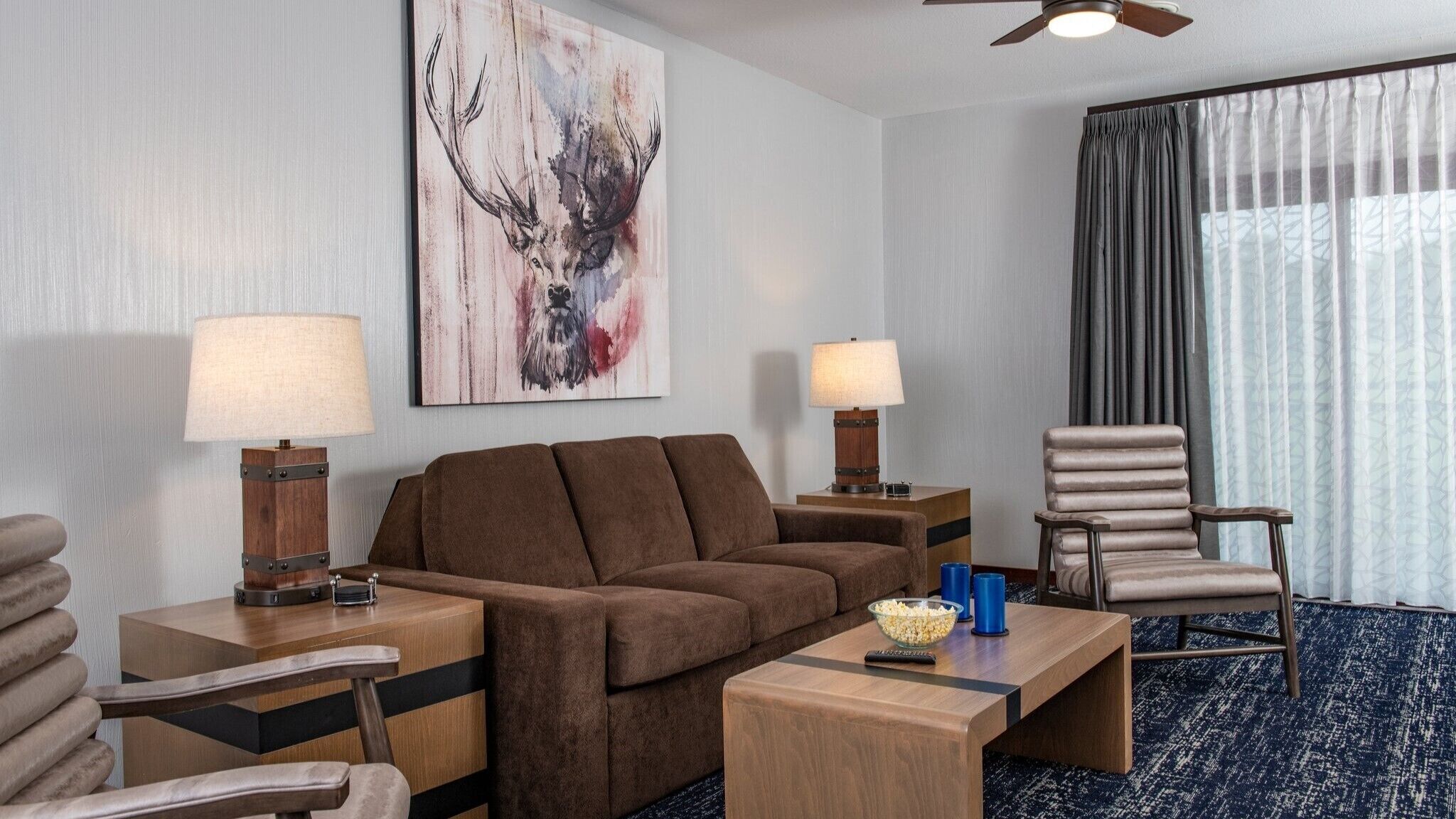 Une chambre d’hôtel avec un canapé marron, deux tables d’appoint avec des lampes, une table basse en bois, une télécommande et un grand tableau abstrait accroché au mur.
