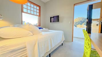 A bedroom with a bed, pillows, a TV on the wall, and a view of buildings through a window.