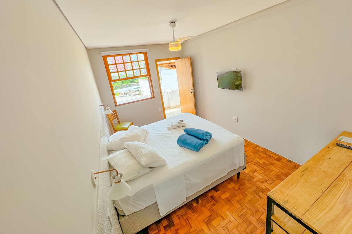 A bedroom with a bed, a wooden table, a TV, and a window with a view of buildings.