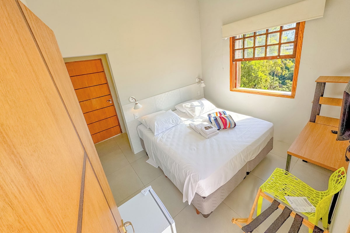 A bedroom with a bed, a window with a view of greenery, a wooden door, and a small table with a chair.