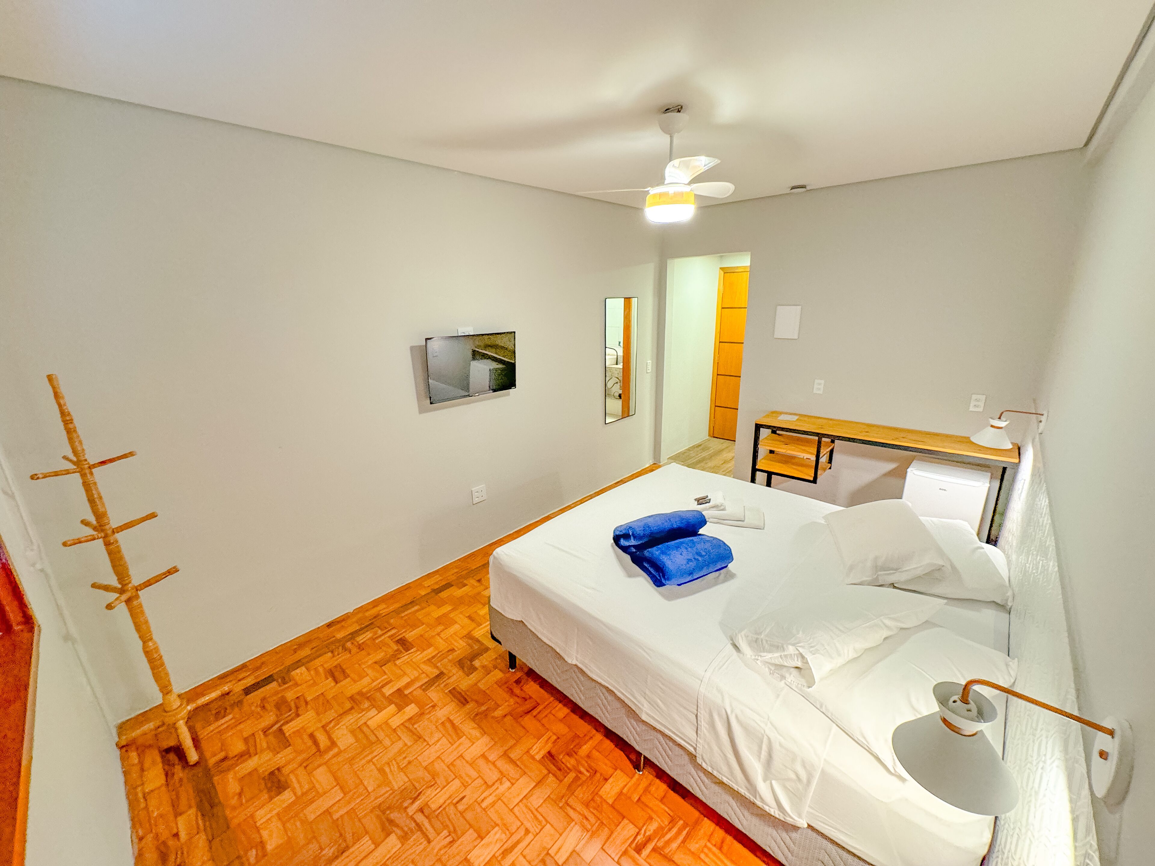 Une chambre avec un plancher en bois, un lit avec une literie blanche, un ventilateur de plafond, une petite table de chevet avec une lampe et un miroir au mur.