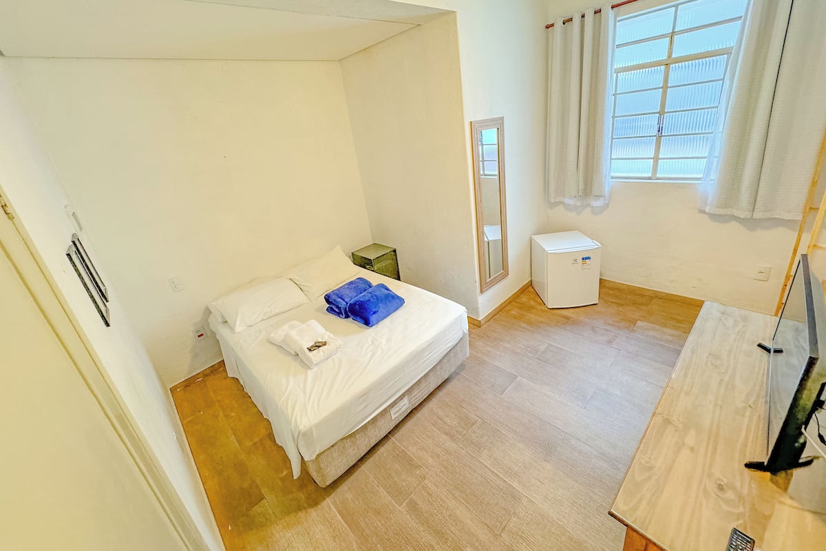 A small, bright room with a bed, a small white heater, a window with curtains, and a wooden floor.