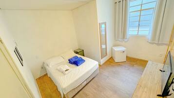 A small, bright room with a bed, a small white heater, a window with curtains, and a wooden floor.