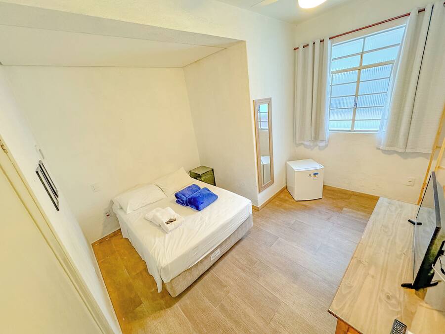 A small, bright room with a bed, a small white heater, a window with curtains, and a wooden floor.