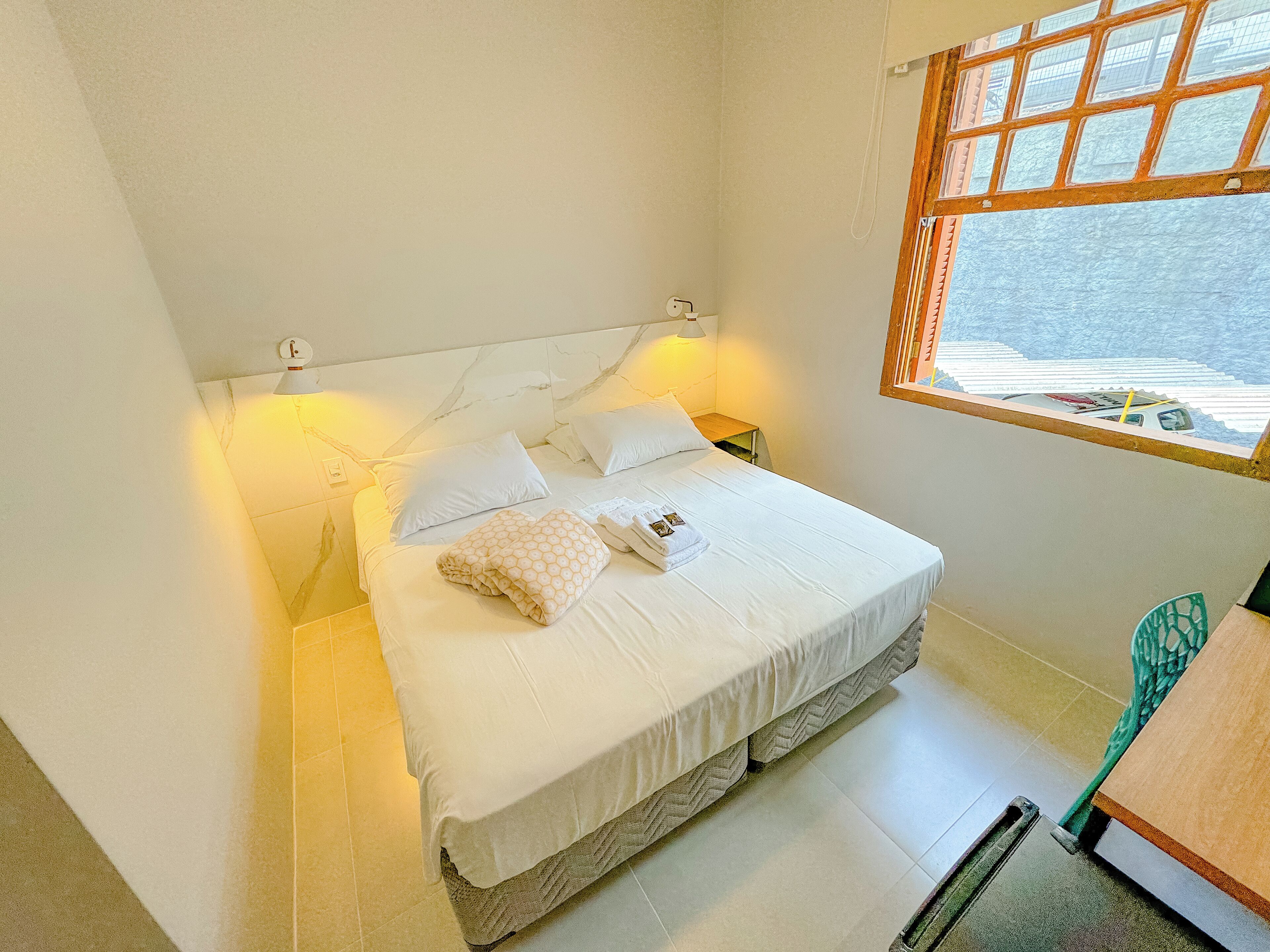 A neatly made bed with pillows and a folded blanket, a bedside table with a lamp, and a window with a view of the outside.