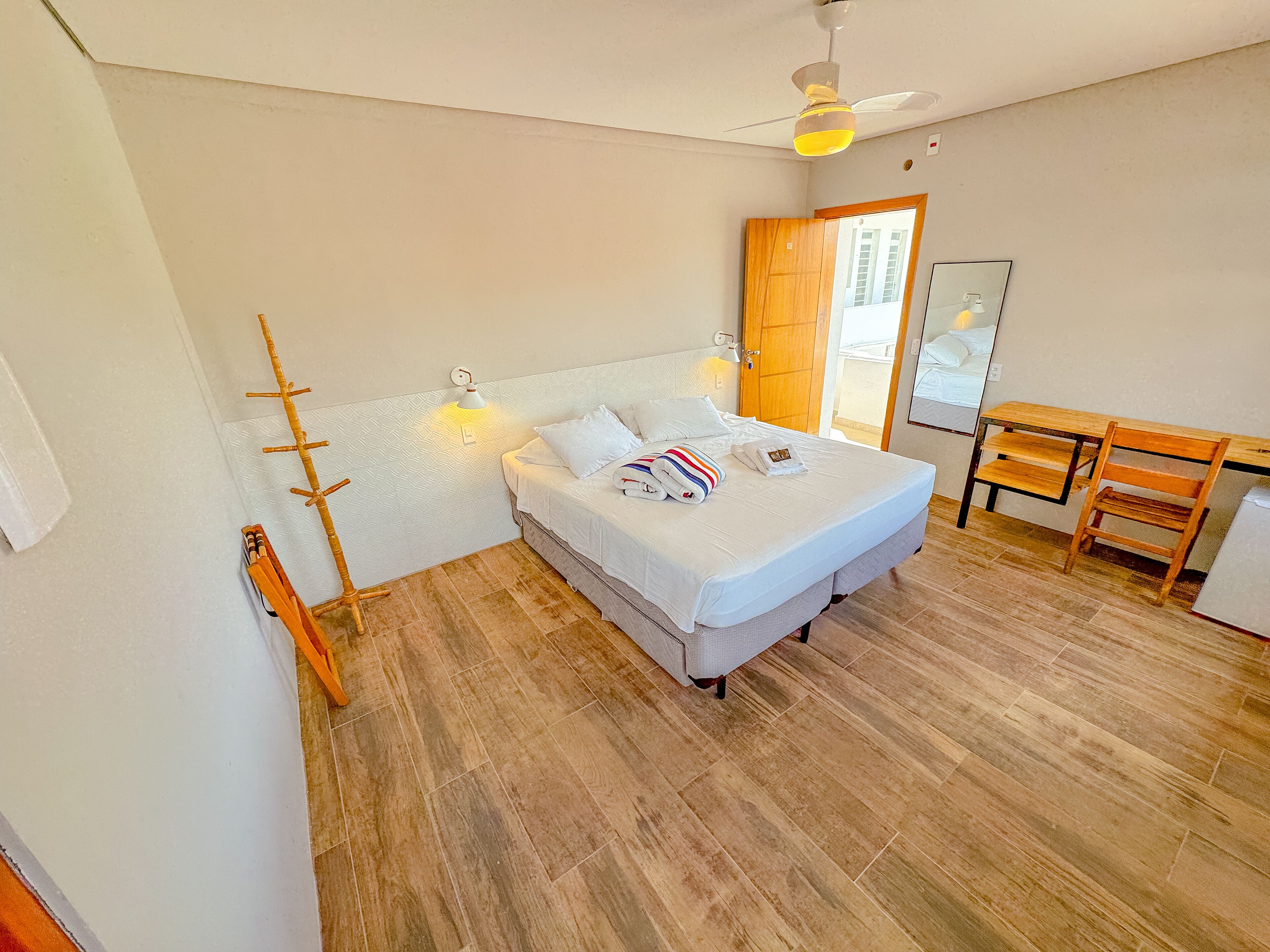 Quarto com cama, escrivaninha, cadeira, piso de madeira, ventilador de teto e porta que dá para outro cômodo.