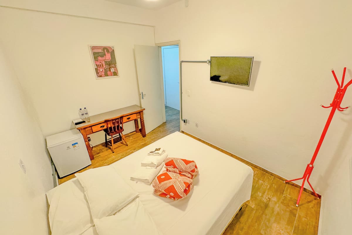 A bedroom with a bed, a desk, a chair, a small fridge, and a red cleaning tool.