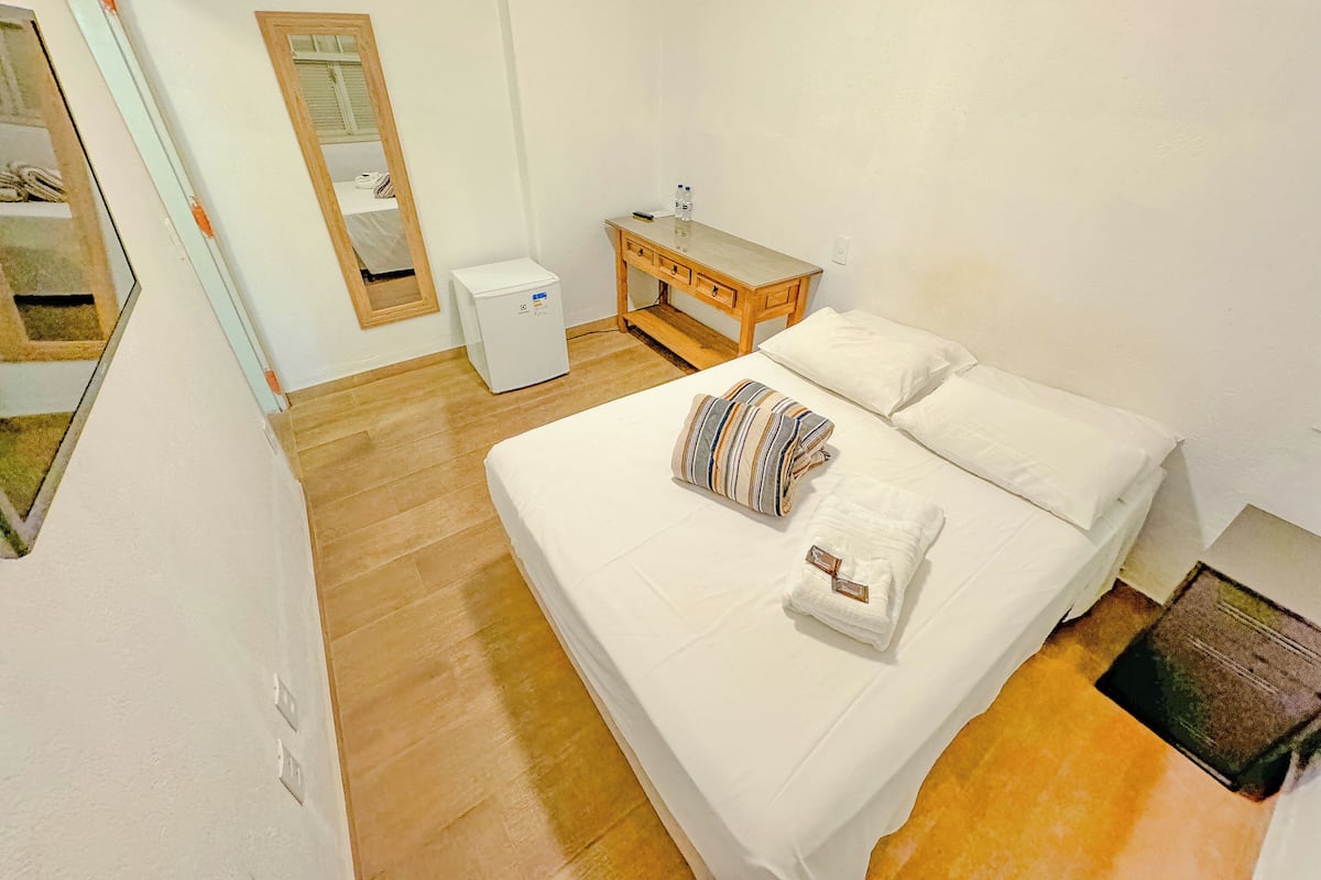 A bedroom with a bed, a wooden nightstand, a small white refrigerator, a mirror, and a wooden floor.
