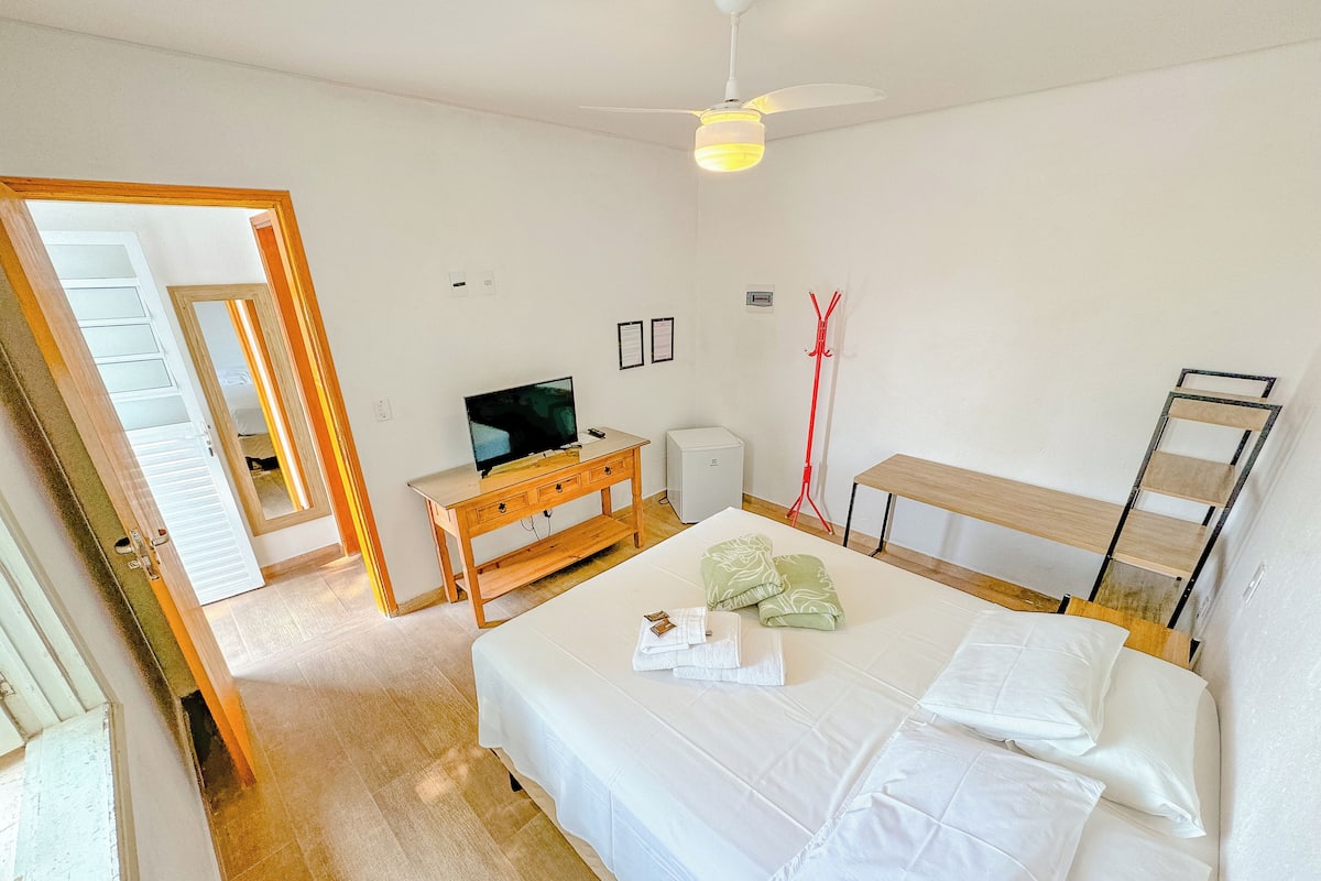 A bedroom with a bed, a TV on a wooden stand, a desk, and a ladder against the wall.