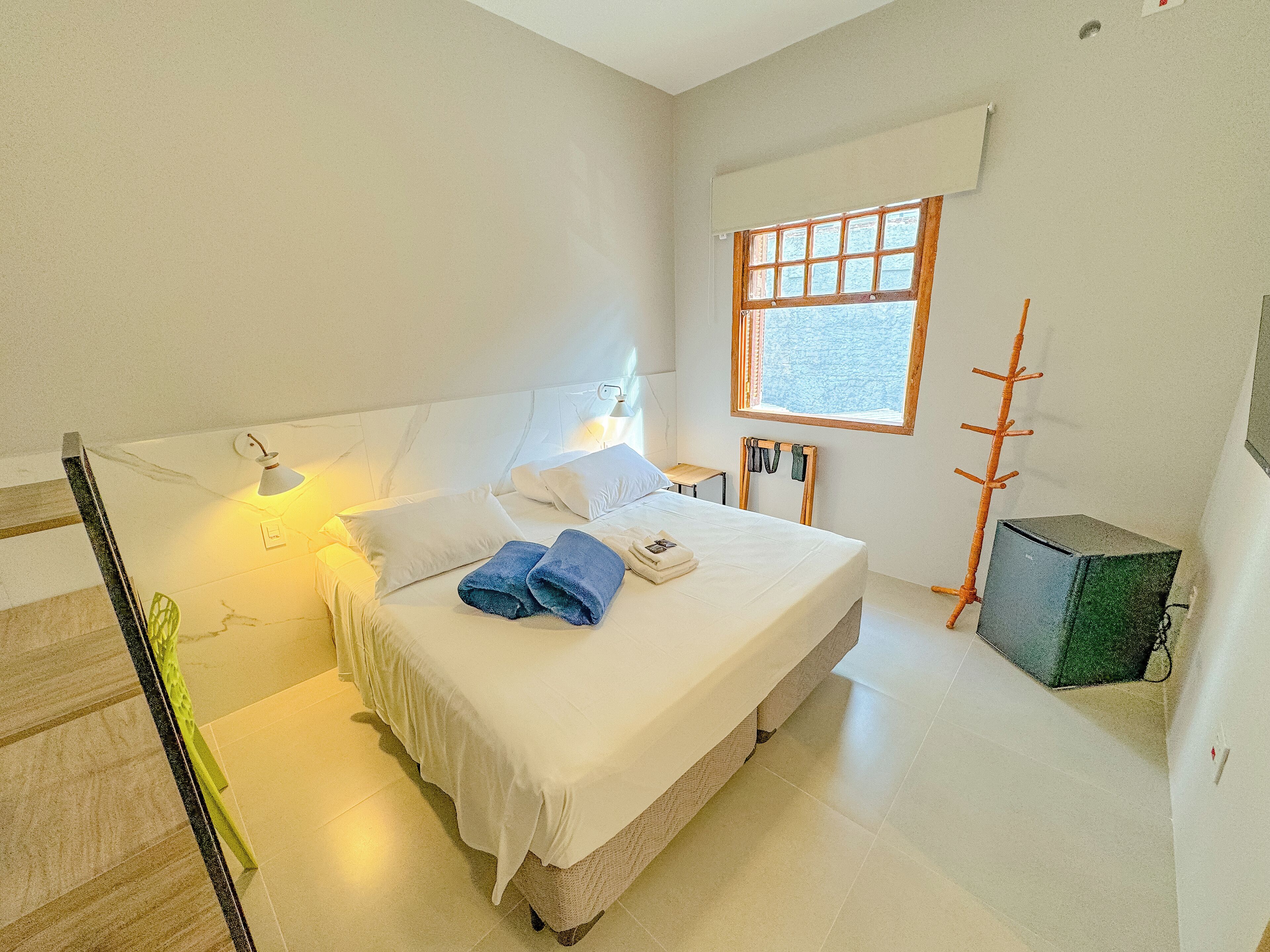 A bedroom with a bed, pillows, a lamp, a window, and a coat rack.