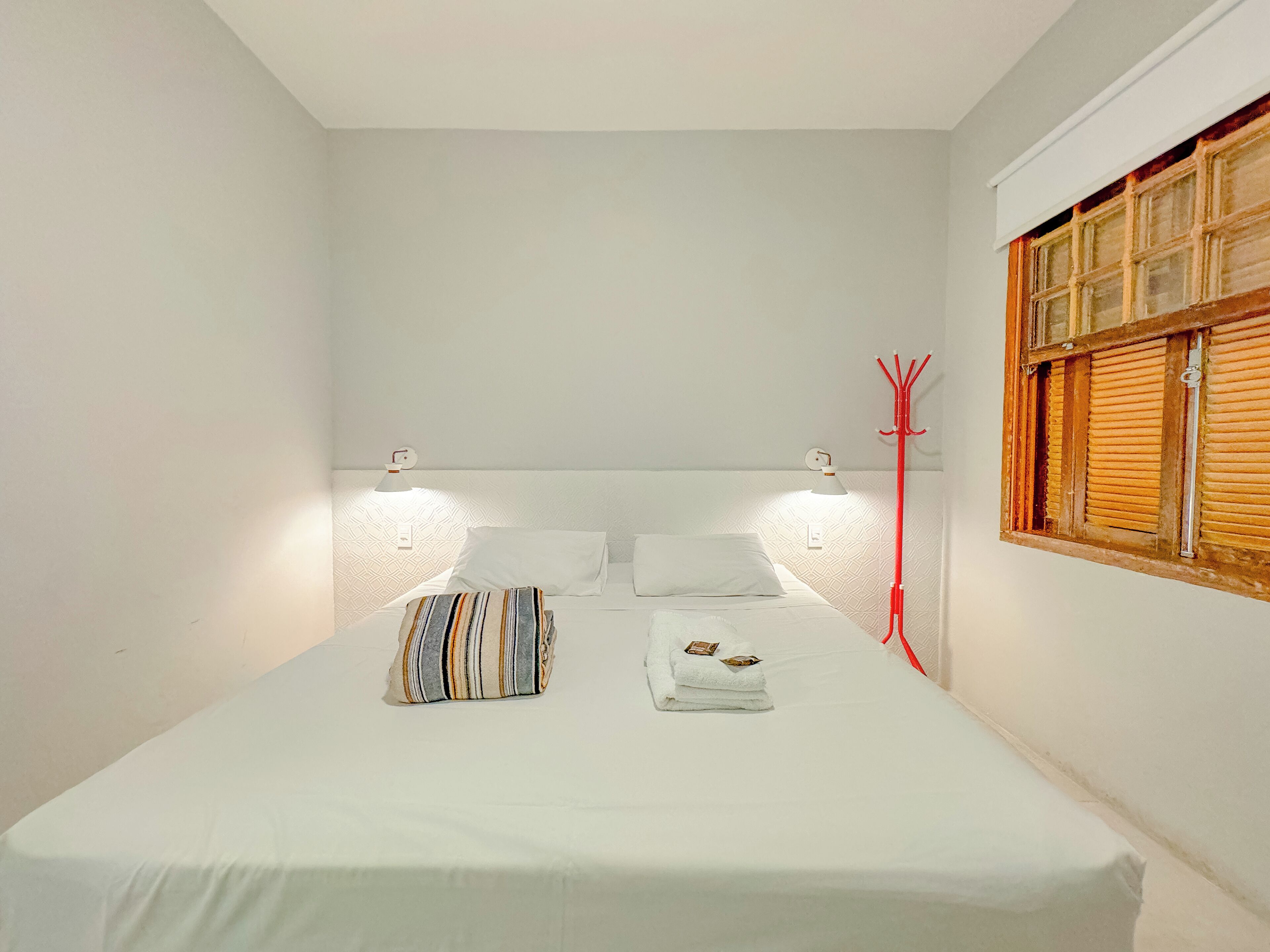Un dormitorio con una cama, dos lámparas de noche, un cojín a rayas y una ventana con contraventanas de madera.
