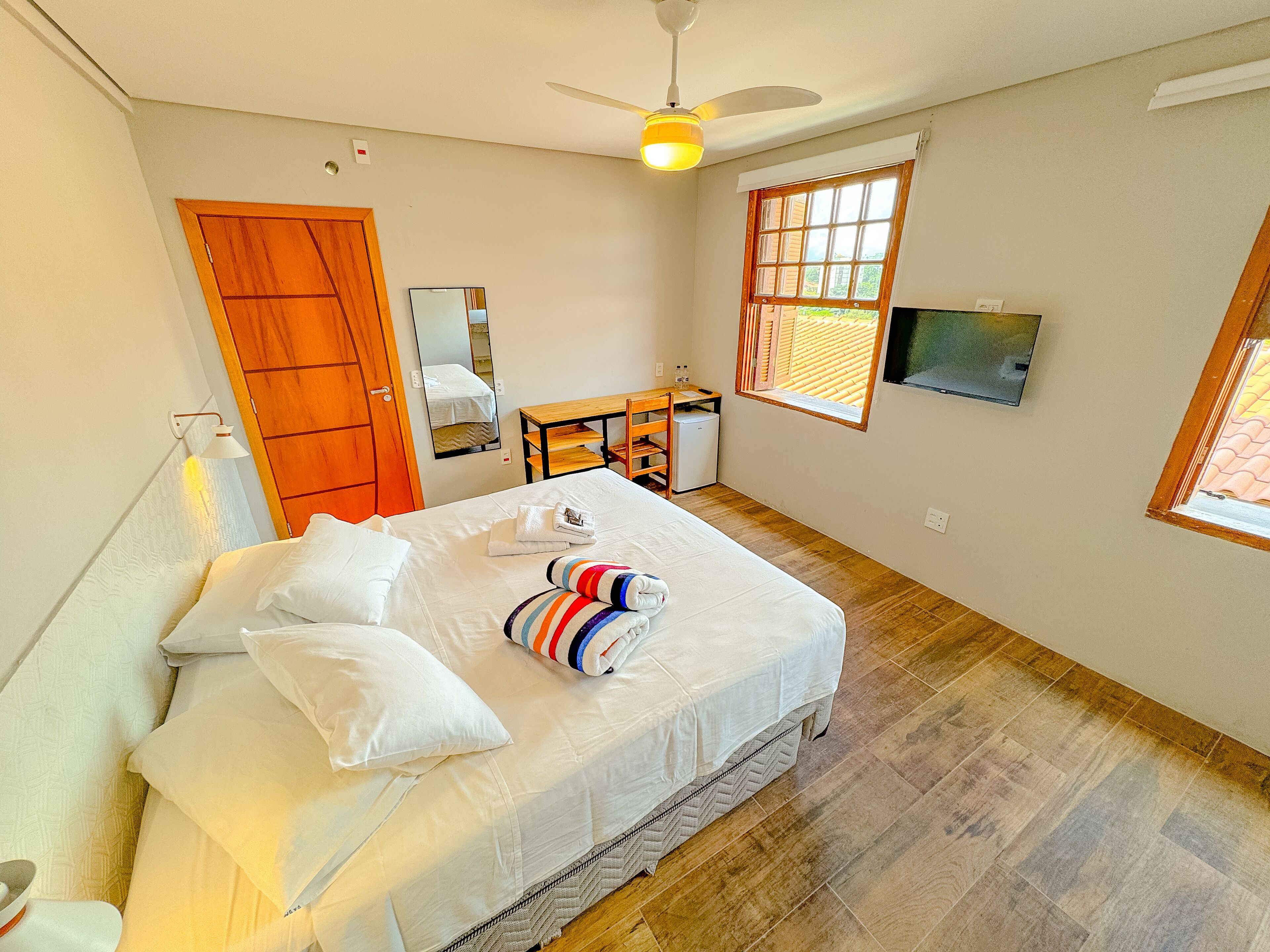 A bedroom with a bed, a desk, a chair, a small refrigerator, and a window.