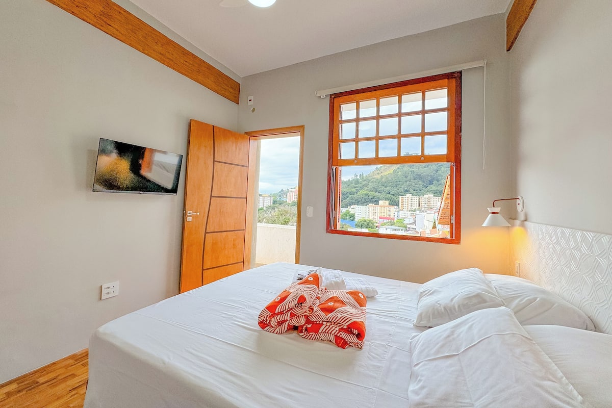 A bedroom with a bed, a window with a view of buildings, a door, and a wall-mounted TV.