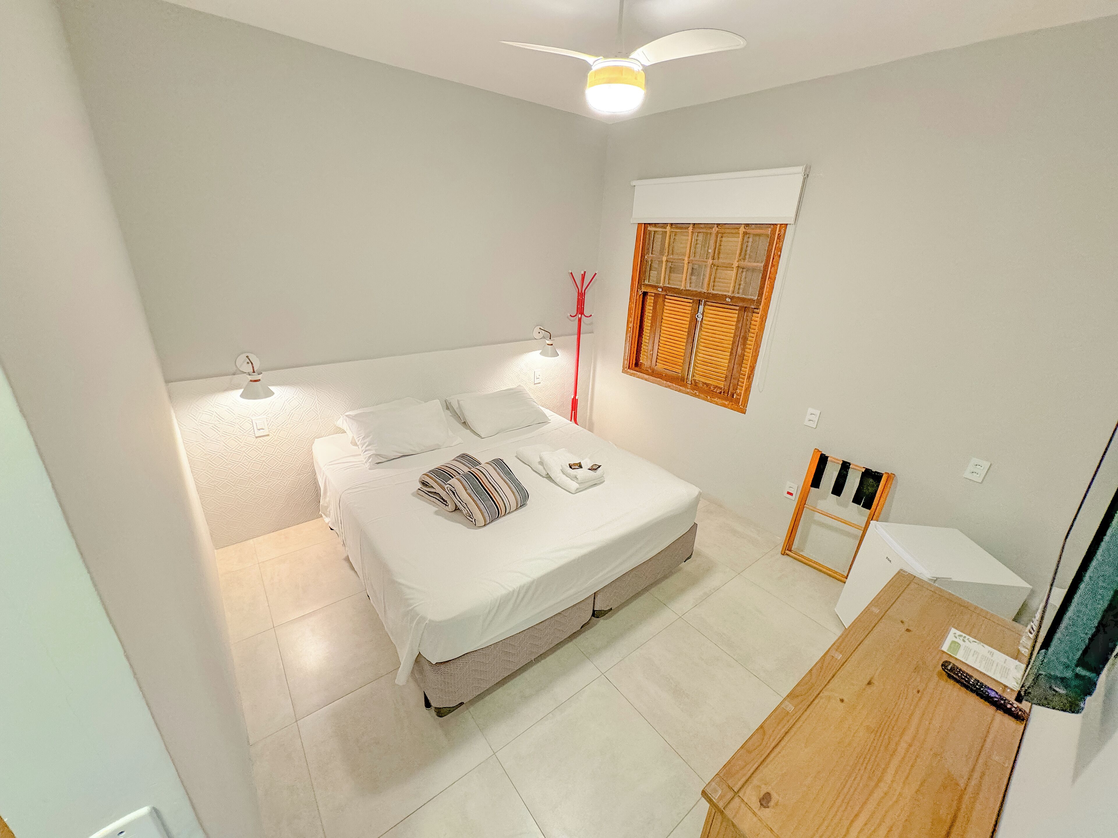 A bedroom with a bed, bedside lamps, a window with shutters, a ceiling fan, and a wooden table.