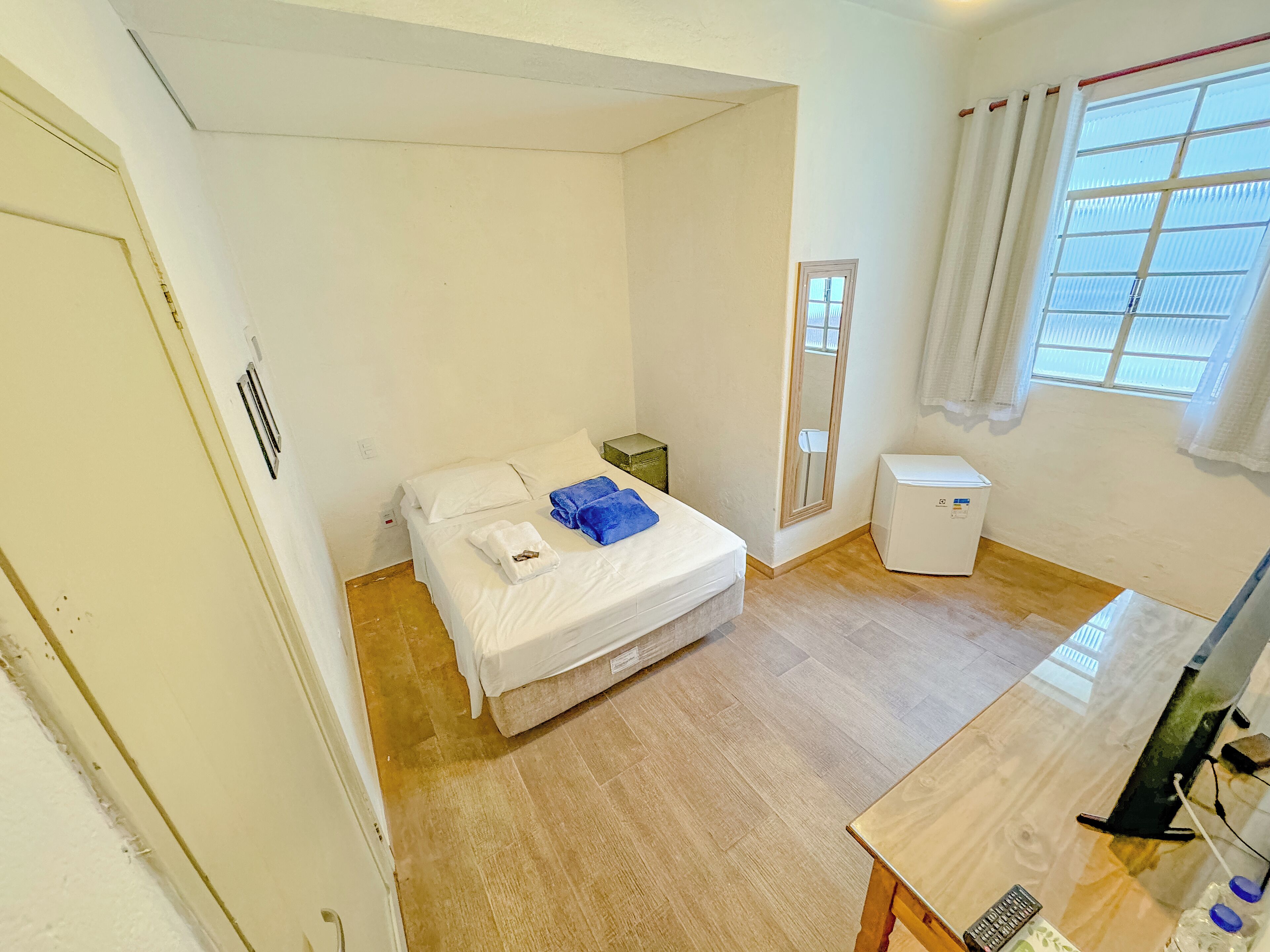 Una habitación pequeña y ordenada con una cama individual, una mesita de noche, un armario blanco pequeño y una ventana con cortinas.