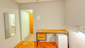 A small, compact room with a wooden desk, a white refrigerator, and a mirror on the wall. - Edifício Barros (Serra Negra)