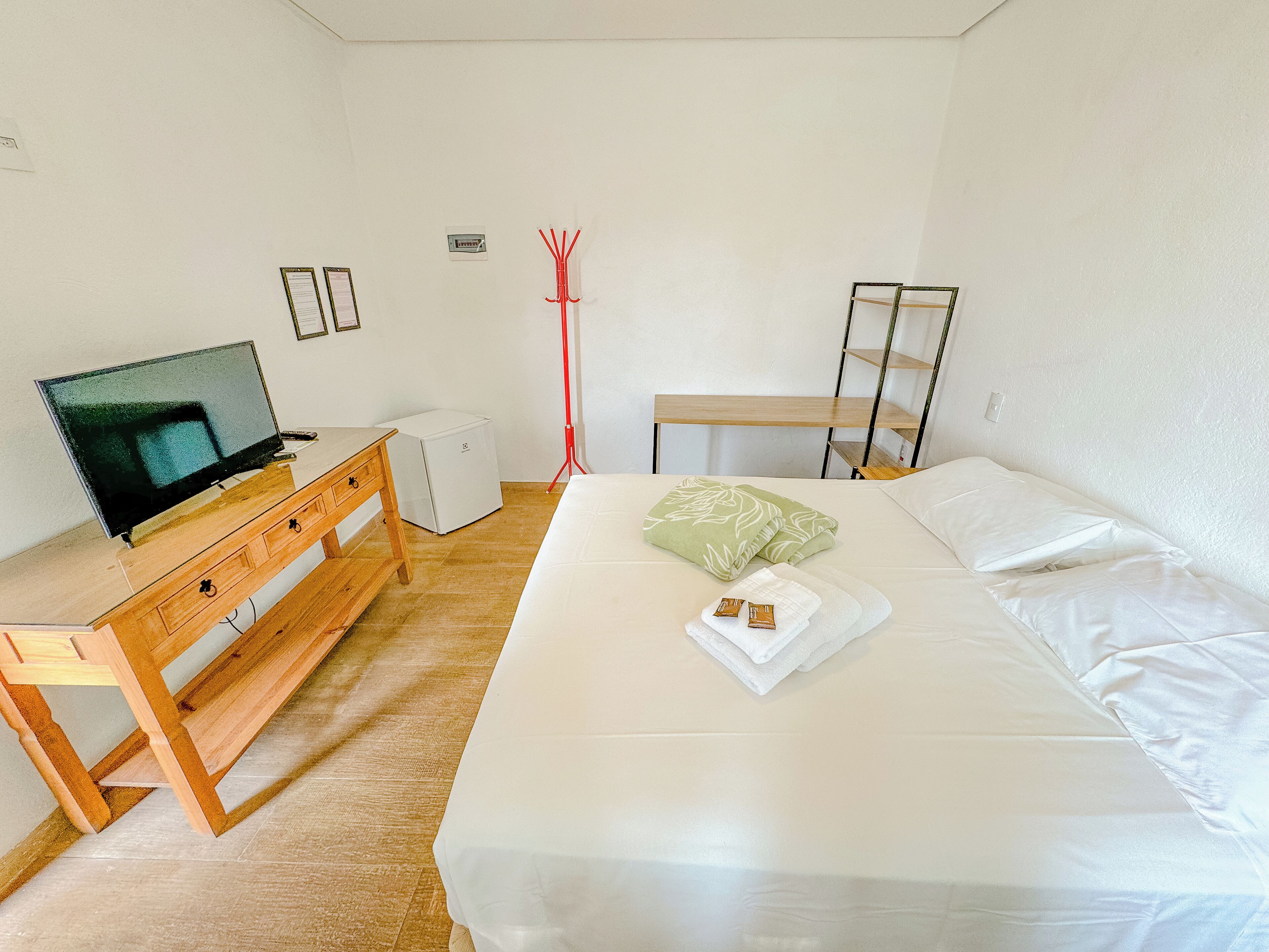 Quarto com cama, televisão sobre uma cômoda de madeira, uma pequena geladeira branca e uma escrivaninha de madeira com cadeira.