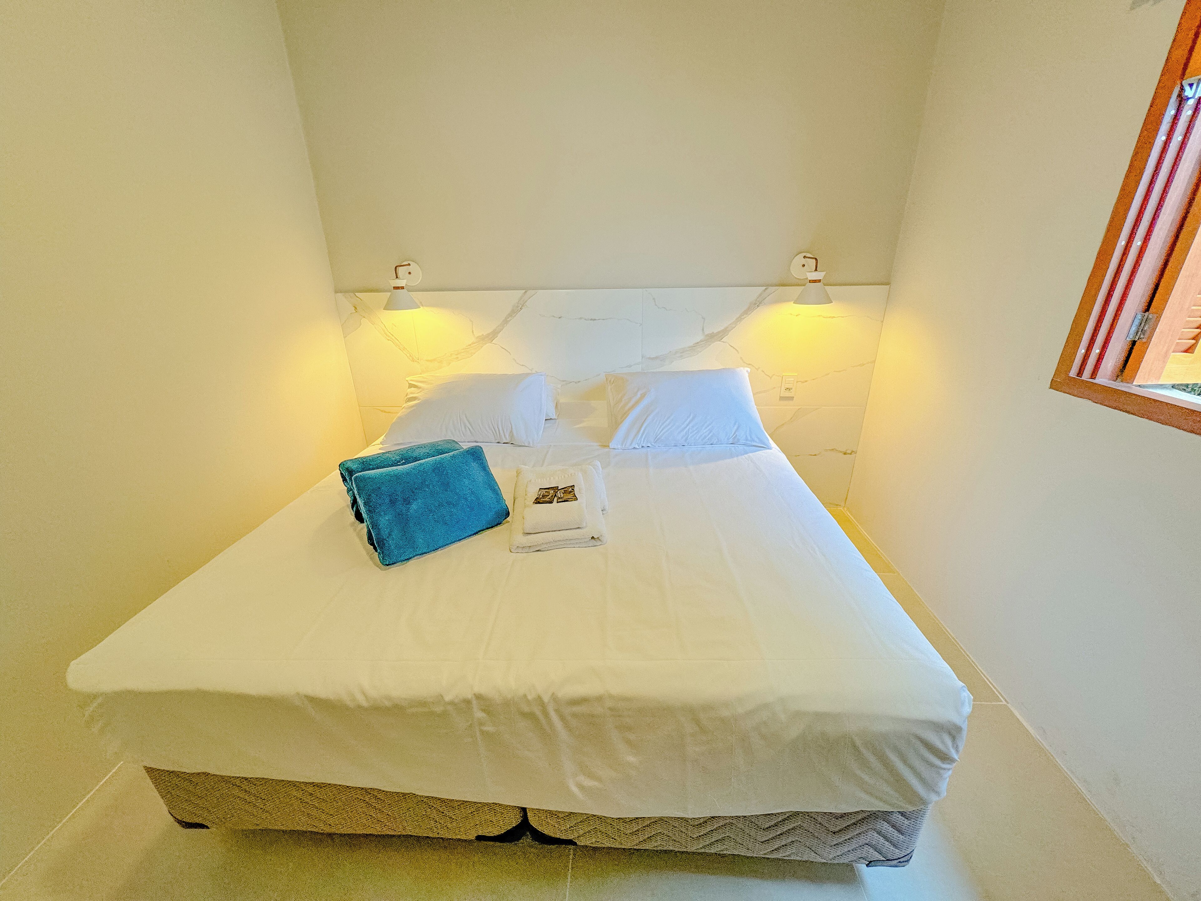 Een net opgemaakt bed met witte lakens en een blauwe handdoek, twee witte kussens, en een klein boekje op het bed.