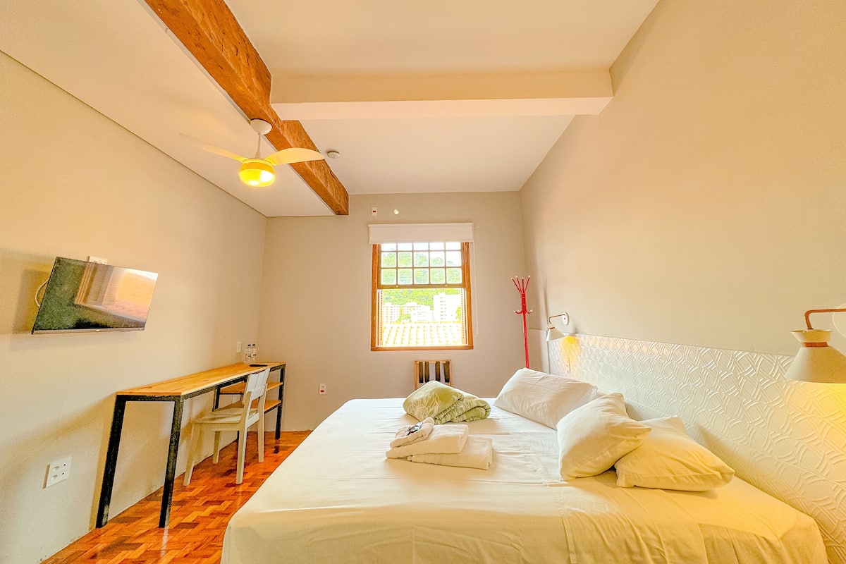A bedroom with a bed, a desk, and a chair. There is a window with a view of greenery outside.