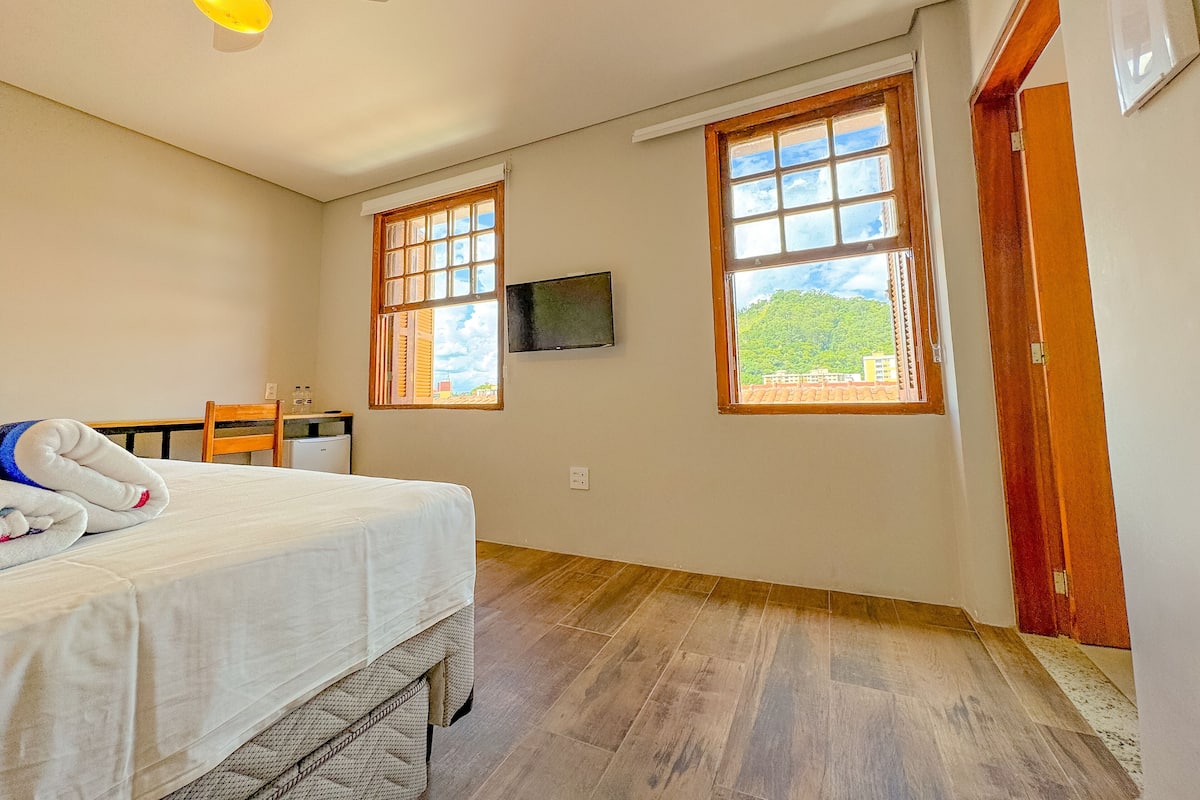 A room with a bed, a desk, two windows, a ceiling fan, and a television.