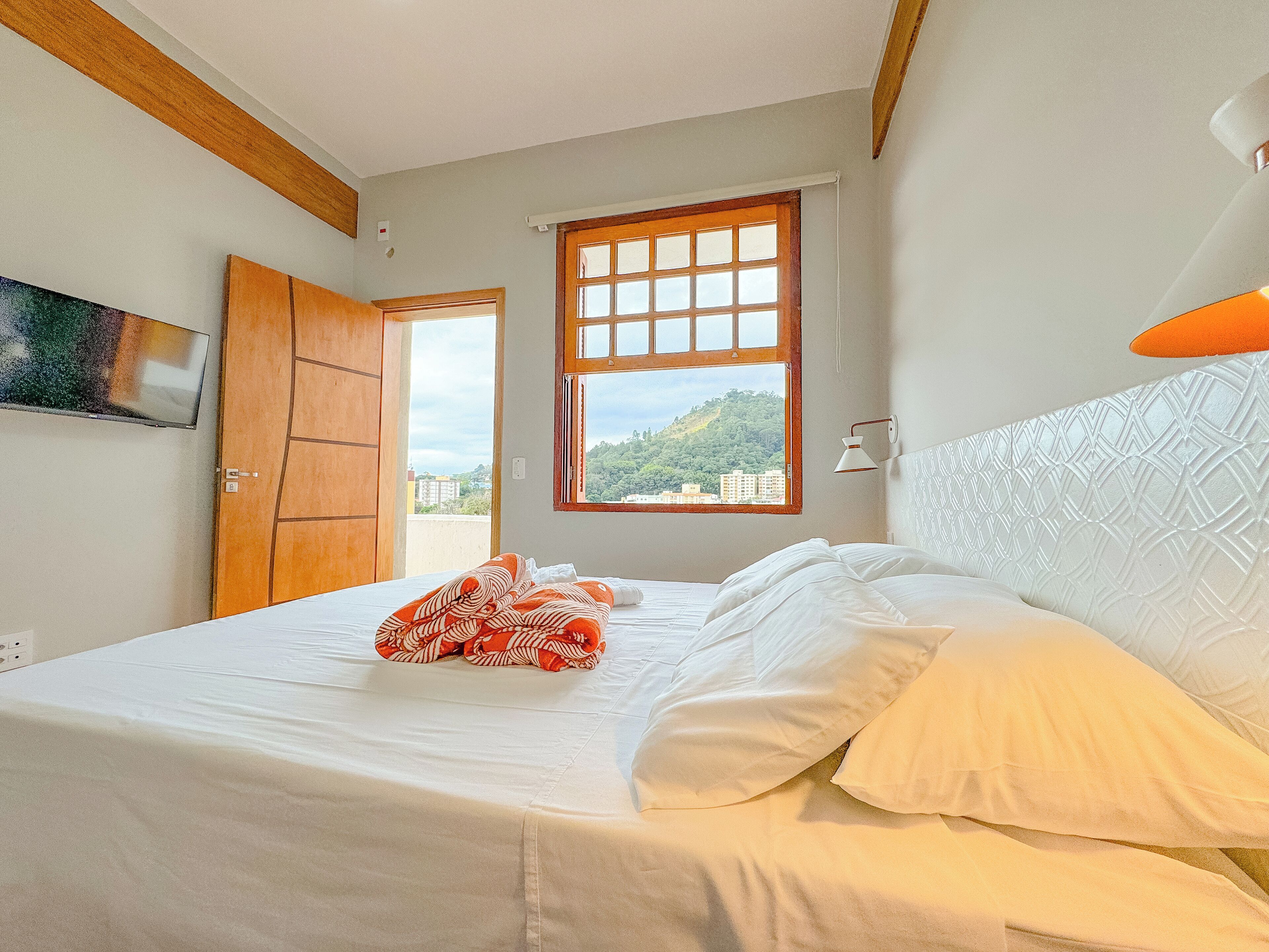 A bedroom with a bed, a window with a view of a cityscape, a wooden door, and a wall-mounted TV.