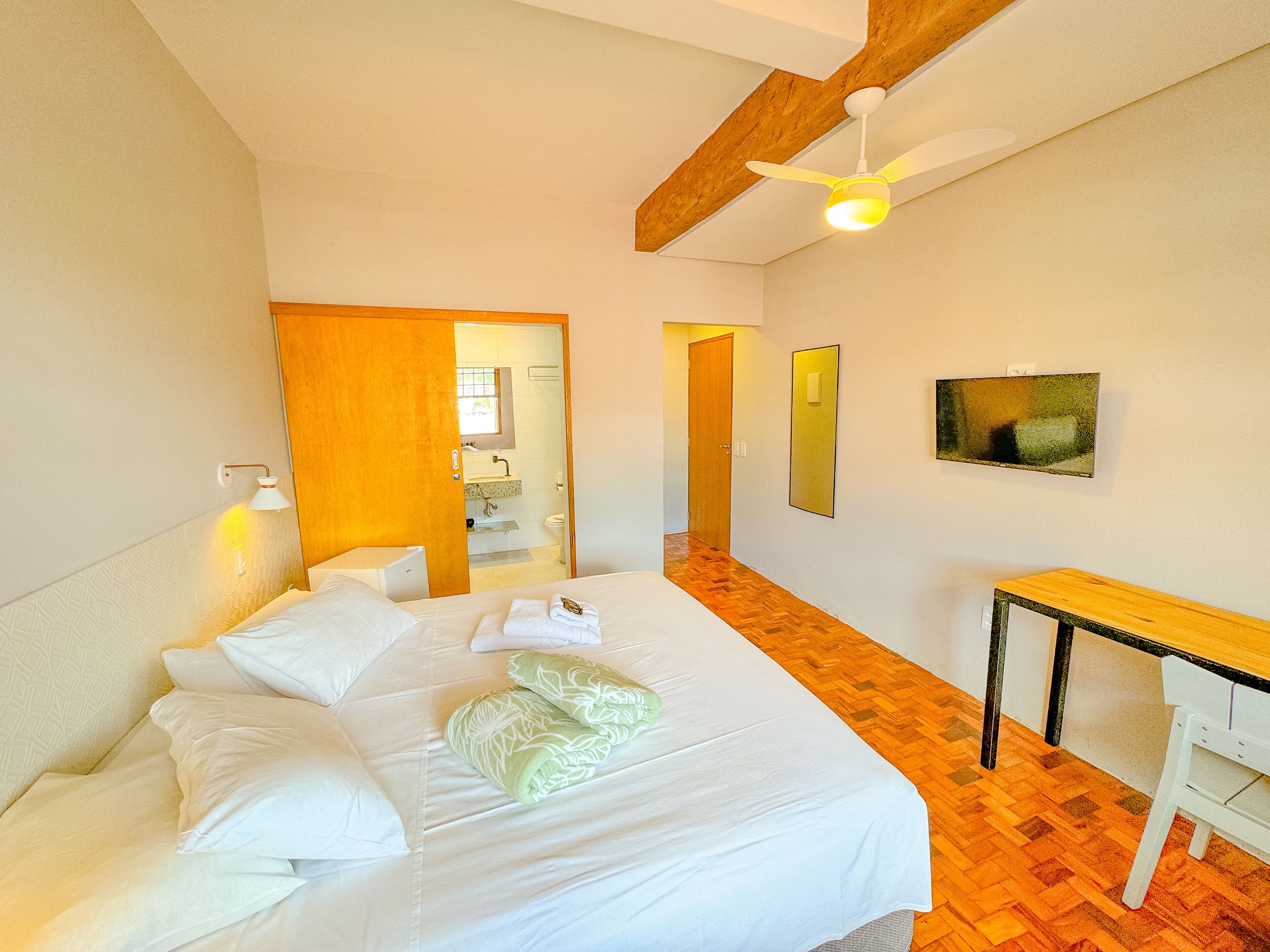 Quarto de hotel com cama, escrivaninha, espelho e ventoinha no teto.